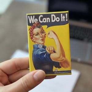 May include: Rectangular magnet featuring the iconic Rosie the Riveter image. The image shows a woman flexing her arm with the text "We Can Do It!" in a speech bubble. The background is yellow, and the woman wears a blue shirt and a red and white polka dot bandana.