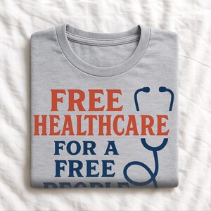 May include: A gray t-shirt with the words "FREE HEALTHCARE FOR A FREE" in red and blue block letters. A blue stethoscope graphic is on the right side of the text. The shirt is folded on a white surface.