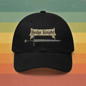 May include: Black baseball cap with the embroidered text "Hedge Knight" above a sword design. The cap has a curved brim and a fabric strap closure on the back. The design is in shades of tan and silver.