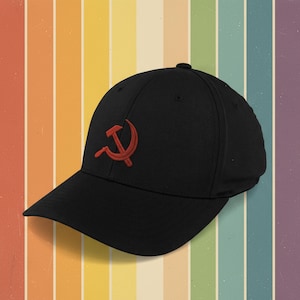 May include: A black baseball cap with a red embroidered hammer and sickle design. The cap has a curved peak and a button on the crown. The background is a striped pattern in shades of orange, yellow, and purple.