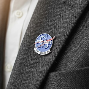 Könnte beinhalten: Runder, blau-weißer Emaille-Anstecker mit dem Text "NOT BEAT" und "WE CHECKED" in weißer Schrift. Der Anstecker hat ein rotes Swoosh-Design und ist an einem grauen Blazer befestigt.
