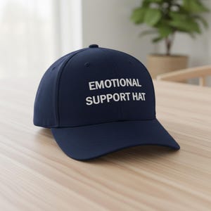 May include: A navy blue baseball cap with the text "EMOTIONAL SUPPORT HAT" in white block letters. The hat is sitting on a light-colored wooden surface. The background includes a window and a potted plant.