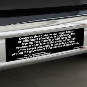 Op de afbeelding: Een zwarte rechthoekige bumpersticker met witte tekst. De tekst is het Eerste Amendement van de Amerikaanse grondwet, dat de vrijheid van godsdienst, meningsuiting, pers, vergadering en het recht op petitie garandeert.