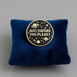 May include: A circular, black and gold enamel pin featuring the words "JUST VISITING THIS PLANET" and a space design. The pin is displayed on a small, dark blue velvet cushion.