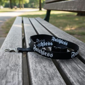 May include: Black lanyard with the word "Faithless" in white gothic font. The lanyard has a silver-colored metal clasp and a black plastic buckle. The lanyard is lying on a wooden bench.