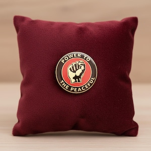 Könnte beinhalten: Ein runder Ansteck-Pin mit dem Schriftzug „POWER TO THE PEACEFUL“, der ein erhobenes Faust-Design umgibt. Der Pin liegt auf einem weinroten Samtkissen.