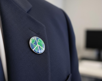 May include: A round, silver-toned pin featuring a blue and green world map design with a white peace symbol overlay. The pin is affixed to a navy blue blazer, suggesting a theme of global peace and environmental awareness.