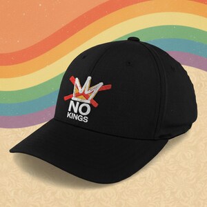 Peut inclure: Casquette de baseball noire avec un motif brodé. Le motif représente une couronne barrée avec les mots "NO KINGS" en blanc. L'arrière-plan présente un dégradé arc-en-ciel.