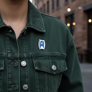 May include: A blue and white enamel pin in a geometric design is affixed to a dark green denim jacket. The pin is a stylized shape with a blue center and white border, creating a modern aesthetic. The jacket has silver buttons.