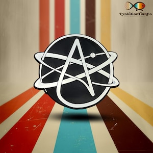 Op de afbeelding: Een zwart-witte emaille pin met het symbool van een 'A' in een cirkel met een gestileerd atoomontwerp. De pin is op een gestreepte achtergrond.