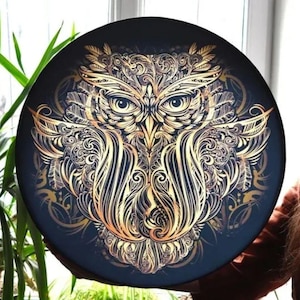 Puede incluir: Un plato decorativo redondo y negro con un diseño detallado de búho dorado. La cara del búho está intrincadamente estampada con plumas y líneas onduladas. El plato se sostiene frente a una planta verde.