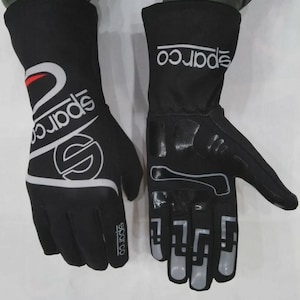 May include: Pair of black Sparco racing gloves. The gloves feature white and red accents, with the Sparco logo prominently displayed. The design includes reinforced areas on the palm and fingers for enhanced grip and protection. The gloves are designed for motorsports.