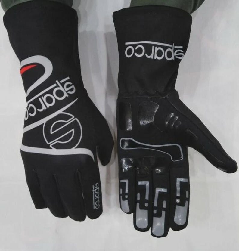 May include: Pair of black Sparco racing gloves. The gloves feature white and red accents, with the Sparco logo prominently displayed. The design includes reinforced areas on the palm and fingers for enhanced grip and protection. The gloves are designed for motorsports.