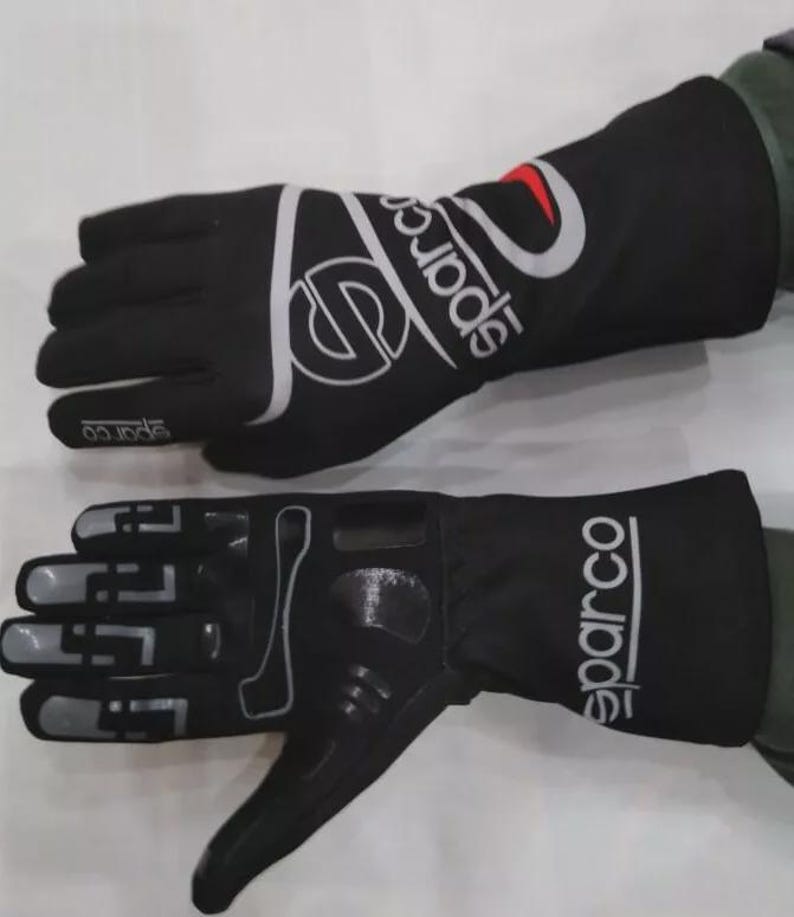 May include: Pair of black Sparco racing gloves. The gloves have a white and silver Sparco logo on the back of the hand and the wrist. The palm of the glove has a textured grip. The gloves are designed for motorsports.