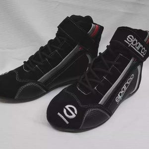 May include: Pair of black Sparco racing shoes. The high-top shoes have a suede-like texture, white stitching, and a velcro strap. The Sparco logo is visible on the strap and side of the shoe. The toe of the shoe has a white graphic.