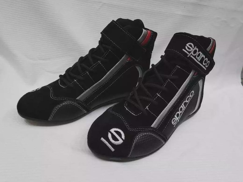 May include: Pair of black Sparco racing shoes. The high-top shoes have a suede-like texture, white stitching, and a velcro strap. The Sparco logo is visible on the strap and side of the shoe. The toe of the shoe has a white graphic.
