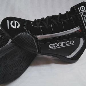May include: Black racing shoes with the Sparco logo in white. The shoes have a black and silver design with white stitching. The sole is black with a textured pattern. The shoes have a velcro strap with the Sparco logo.