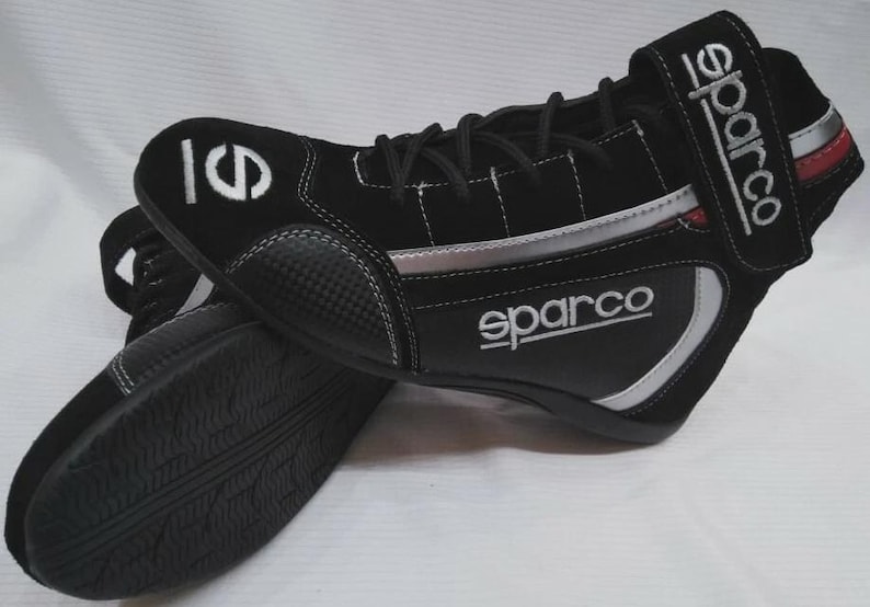 May include: Black racing shoes with the Sparco logo in white. The shoes have a black and silver design with white stitching. The sole is black with a textured pattern. The shoes have a velcro strap with the Sparco logo.