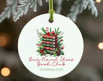 May include: White ceramic Christmas ornament with a green ribbon. The ornament features a stack of books with a red bow, surrounded by holly and pine. Text reads "Our Novel Ideas Book Club Christmas 2025."