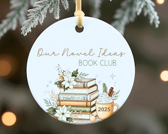 May include: Round Christmas ornament with the text "Our Novel Ideas BOOK CLUB". The ornament features a stack of books, flowers, and a mug with the year 2025. It hangs from a gold ribbon against a backdrop of a snowy tree branch.