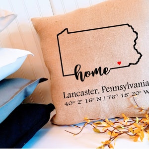 May include: A square burlap pillow with a black outline of Pennsylvania and the word "home" in script. Below, it reads "Lancaster, Pennsylvania" and coordinates. A small red heart marks a location on the map. Other pillows are in the background.