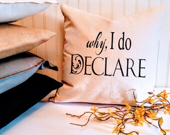 Könnte beinhalten: Ein beiger Kissenbezug mit dem Text "why, I do DECLARE" in schwarzer Schrift. Das Kissen liegt auf einer weißen Oberfläche mit einem Zweig gelber Blumen.
