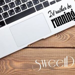 May include: A laptop computer with a black and white keyboard and a white touchpad. A sticker on the laptop says "I'd rather be running." A small potted plant sits to the right. The laptop rests on a wooden surface.