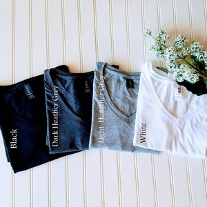 K&ouml;nnte beinhalten: Vier V-Ausschnitt-T-Shirts in verschiedenen Farben: Schwarz, Dunkelgrau meliert, Hellgrau meliert und Wei&szlig;. Jedes Shirt hat ein Etikett mit dem aufgedruckten Farbnamen.