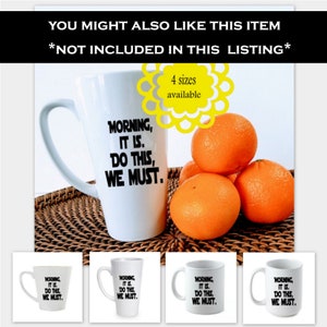 May include: White ceramic mug with the text "Morning, It Is. Do This, We Must." in black lettering. The mug is on a woven brown surface with four oranges in the background.