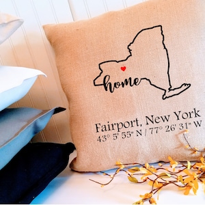 May include: A tan burlap pillow with a black outline of the state of New York and the word "home" in black script. A small red heart is in the center of the state. The text "Fairport, New York 43° 5' 55" N / 77° 26' 31" W" is printed below the state outline.