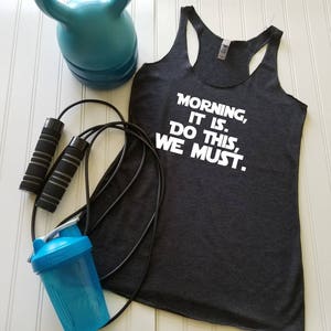 May include: A dark gray tank top with the text "Morning, It Is. Do This, We Must." printed in white. The tank top is paired with a jump rope and a blue shaker bottle.