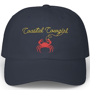 May include: A navy blue baseball cap with the text "Coastal Cowgirl" embroidered in gold thread. A red crab with gold claws is embroidered below the text.