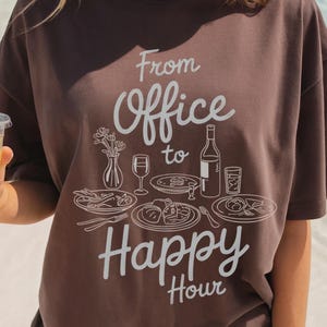 May include: A brown t-shirt with the text "From Office to Happy Hour" in white script. The design includes illustrations of food and drinks. The person is holding a clear plastic cup with a yellow beverage and lime slices.