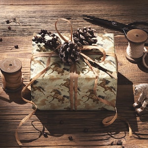 A gift box wrapped in brown paper with a horse pattern sits on a wooden surface, accompanied by a pair of scissors, a spool of thread, and a small toy horse.