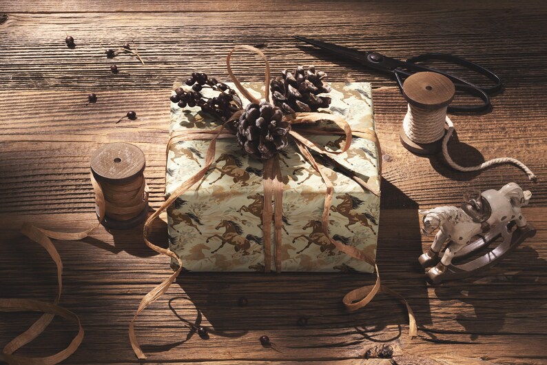 A gift box wrapped in brown paper with a horse pattern sits on a wooden surface, accompanied by a pair of scissors, a spool of thread, and a small toy horse.