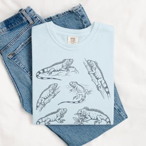 May include: A light blue t-shirt featuring a detailed black and white illustration of several iguanas in various poses. The shirt is folded on top of a pair of blue denim jeans.