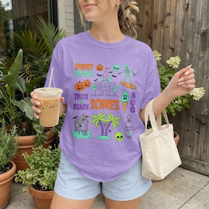 Peut inclure: T-shirt lilas avec un motif graphique de citrouilles, d'un château de sable, d'un squelette sur une planche de surf et les mots "Spooky Summer", "Trick or Beach Scares". Le t-shirt a une coupe décontractée.