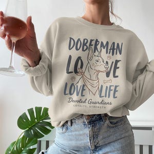 Op de afbeelding: Een beige sweatshirt met de tekst "Doberman Love Life" en een afbeelding van een Dobermann. Het sweatshirt heeft ook de woorden "Devoted Guardians" en "Loyalty, Strength" erop staan.