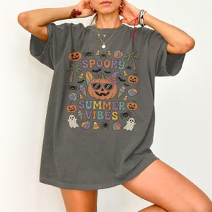 Può includere: T-shirt grigio scuro con una grafica colorata che recita "Spooky Summer Vibes". Il design presenta una zucca con occhiali da sole, circondata da icone a tema Halloween ed estivo come fantasmi, coni gelato e palme.