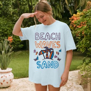 以下が含まれることがあります： 紫とオレンジ色の「BEACH WAVES SAND」の文字が入った水色のTシャツ。傘、サーフボード、ヤシの木が描かれたビーチをテーマにしたグラフィックが特徴です。ゆったりとしたフィット感のTシャツです。