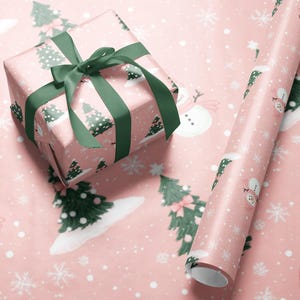 A pink gift box with a green ribbon and a snowman on the wrapping paper, along with a roll of pink and white snowflake-patterned wrapping paper.