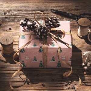 A pink gift box with a Christmas tree pattern and pine cones is wrapped and tied with a ribbon, accompanied by a pair of scissors, a spool of thread, and a small toy horse on a wooden surface.