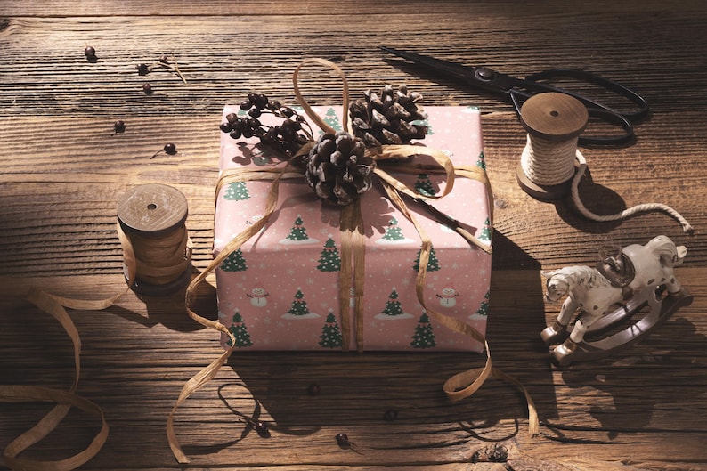 A pink gift box with a Christmas tree pattern and pine cones is wrapped and tied with a ribbon, accompanied by a pair of scissors, a spool of thread, and a small toy horse on a wooden surface.