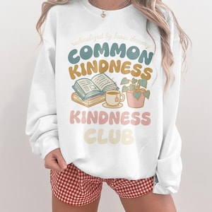 May include: White sweatshirt with the text "radicalized by basic decency COMMON KINDNESS KINDNESS CLUB" in various colors. The graphic includes books, a teacup, and a potted plant. The model is wearing red and white checkered shorts.