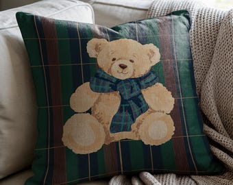 May include: Square decorative pillow featuring a teddy bear wearing a blue and green plaid scarf. The pillow has a green plaid background with brown and navy stripes. The pillow is on a beige couch.