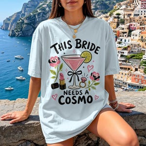 May include: A light blue t-shirt featuring a graphic print with the text THIS BRIDE NEEDS A COSMO. The design includes a pink cocktail in a martini glass with a lime wedge, a lipstick tube, a diamond ring, pink roses, and small hearts.