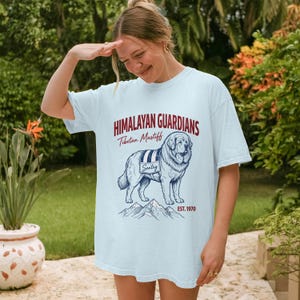 Op de afbeelding: Lichtblauw T-shirt met een afbeelding van een Tibetaanse Mastiff-hond, de tekst "Himalayan Guardians" en "Est. 1970". De hond staat voor een bergketen. Het T-shirt wordt door een persoon gedragen.