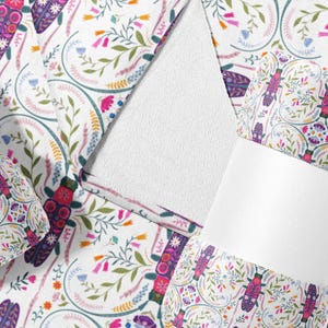 May include: White fabric with a colorful floral and insect pattern. The design includes purple, pink, and orange flowers, along with stylized insects. The fabric is folded and wrapped with white bands, suggesting a textile product.