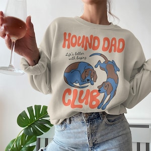 Pode incluir: Camisola bege com o texto laranja "HOUND DAD CLUB". O design apresenta duas ilustrações de cães de caça azuis e castanhos. A frase "Life's better with baying" está impressa abaixo de "HOUND DAD".