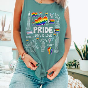 May include: Teal tank top with a white graphic design featuring the word "PRIDE", a rainbow heart, flags, and other symbols. The words "LOVE IS LOVE", "EQUALITY", "CELEBRATE", and "UNITY" are also included in the design.